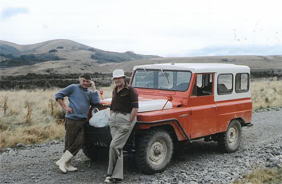 Allan and Jack stand on a gravel track surrounded by tussock grass with farmland hills in the distance. They lean on the front of a red LandRover which as a white roof.