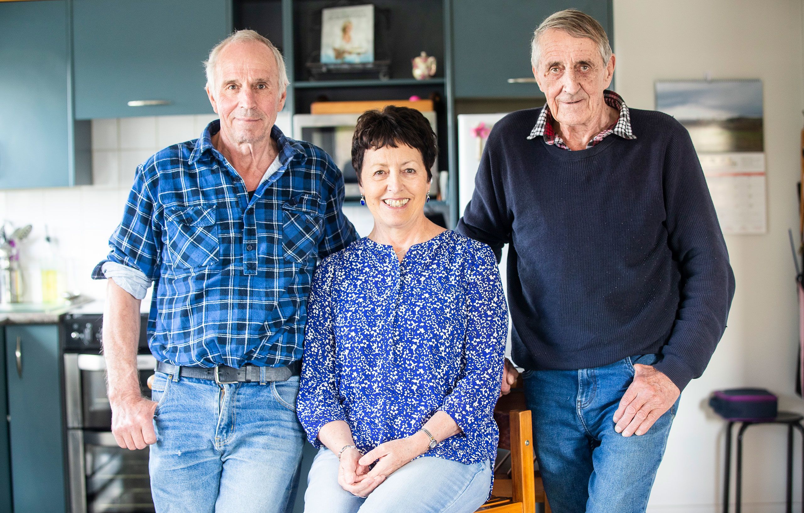 Allan's children, now much older, posing in an open-plan kitchen. They are all looking directly to camera smiling. Mark stands on the left and wears a blue, check patterned, flannel shirt with the sleeves rolled up to his elbows, and faded blue jeans. Kerryn is seated in the middle and wears a blue, ditsy print, long-sleeved blouse and pale blue jeans. Donald stands on the right and wears a dark blue knitted jumper and mid-blue jeans.