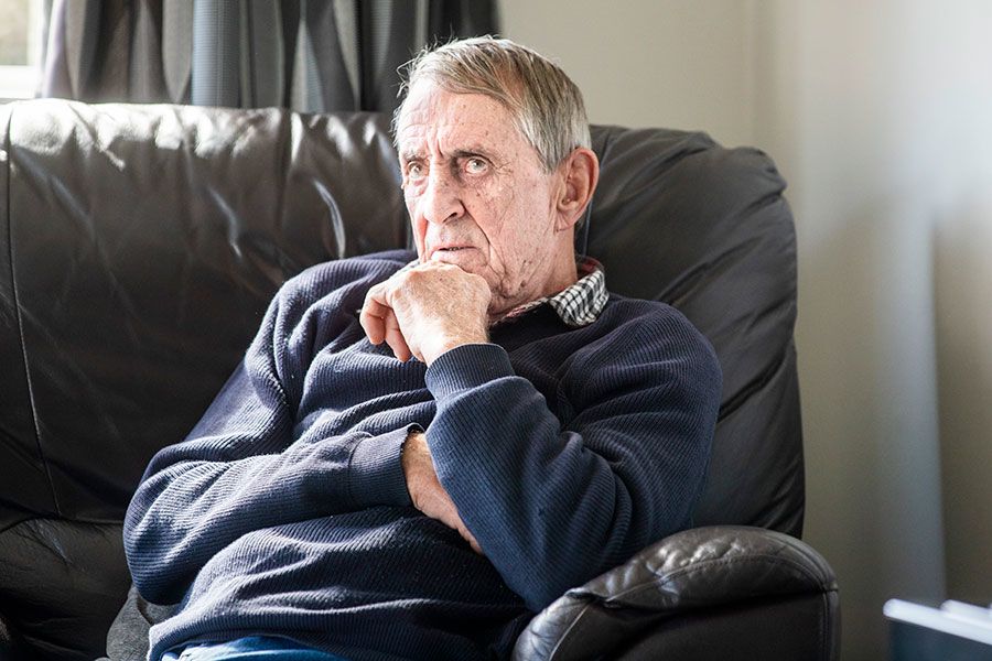 Donald sits on a couch with one arm around his middle and the other hand on his chin. He looks up to the left slightly and has a serious look on his face.
