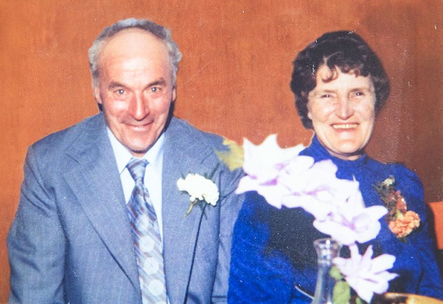 Photograph of Allan and Jean taken at a formal event. Allan wears a silvery blue suit, white shirt and a silvery-blue patterned tie. A white carnation is attached through the buttonhole of his left lapel. Jeans wears a bright blue, long-sleeved top with a high neckline and also wears buttonhole flowers. There is a pink orchid flower arrangement in the foreground.
