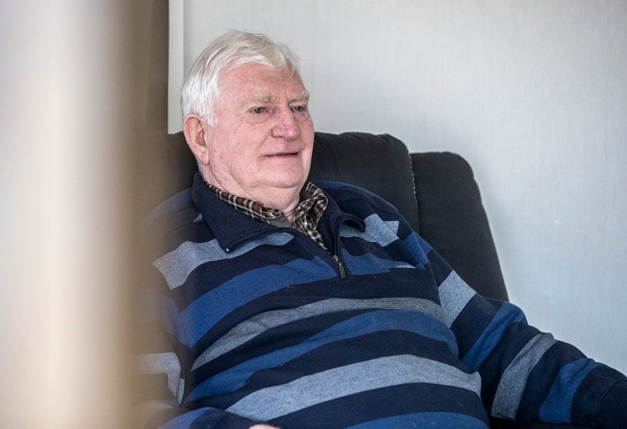 John sits in an armchair, smiling slightly and looking to the right. He wears a blue-striped jumper.