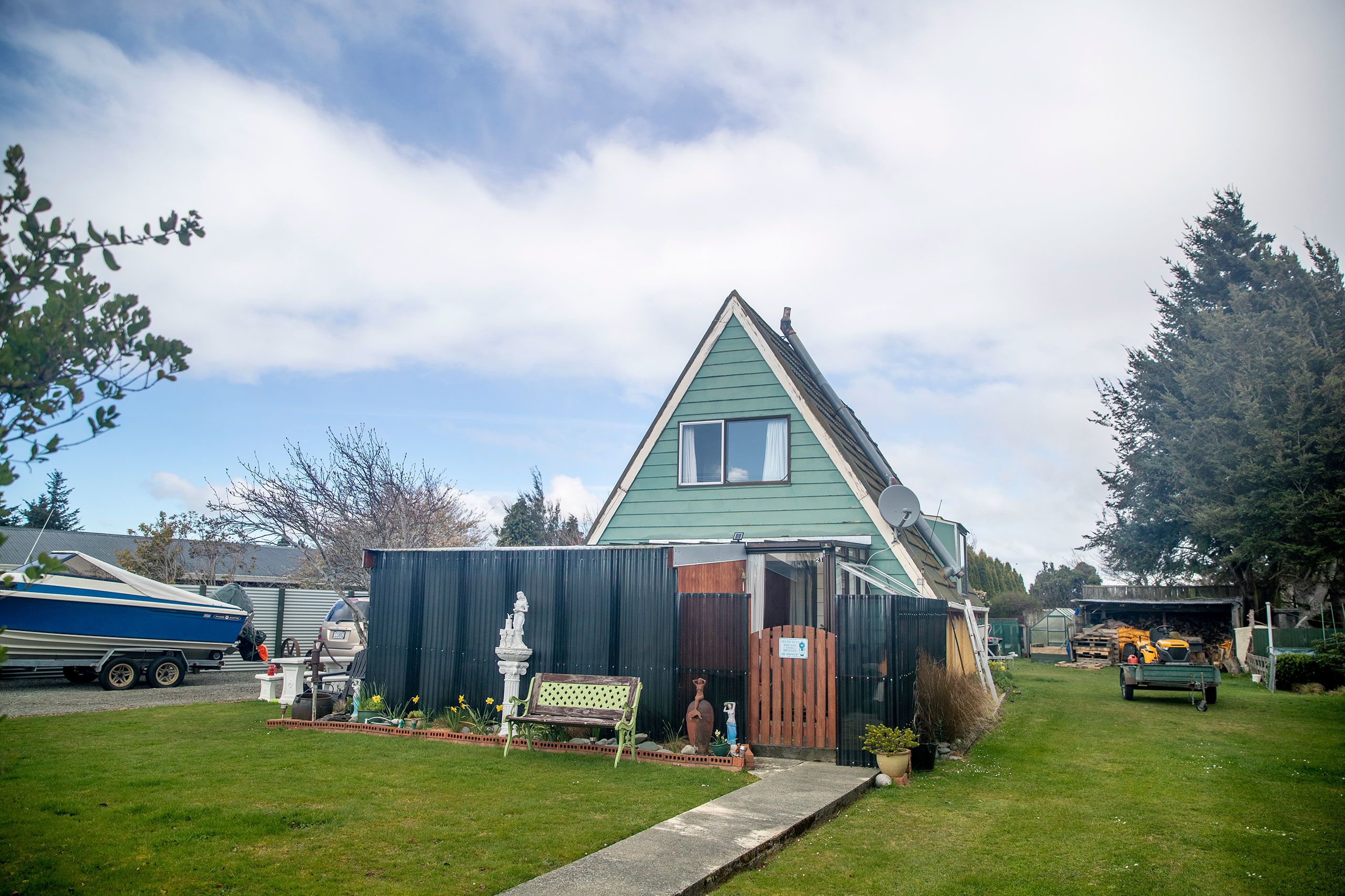 An A-framed weatherboard house painted green with a high, black fence erected at the front before the entrance. The house sits on a large, flat, lawned section. There is a jet boat parked on the driveway to the left and pine trees line the right of the section towards the rear.
