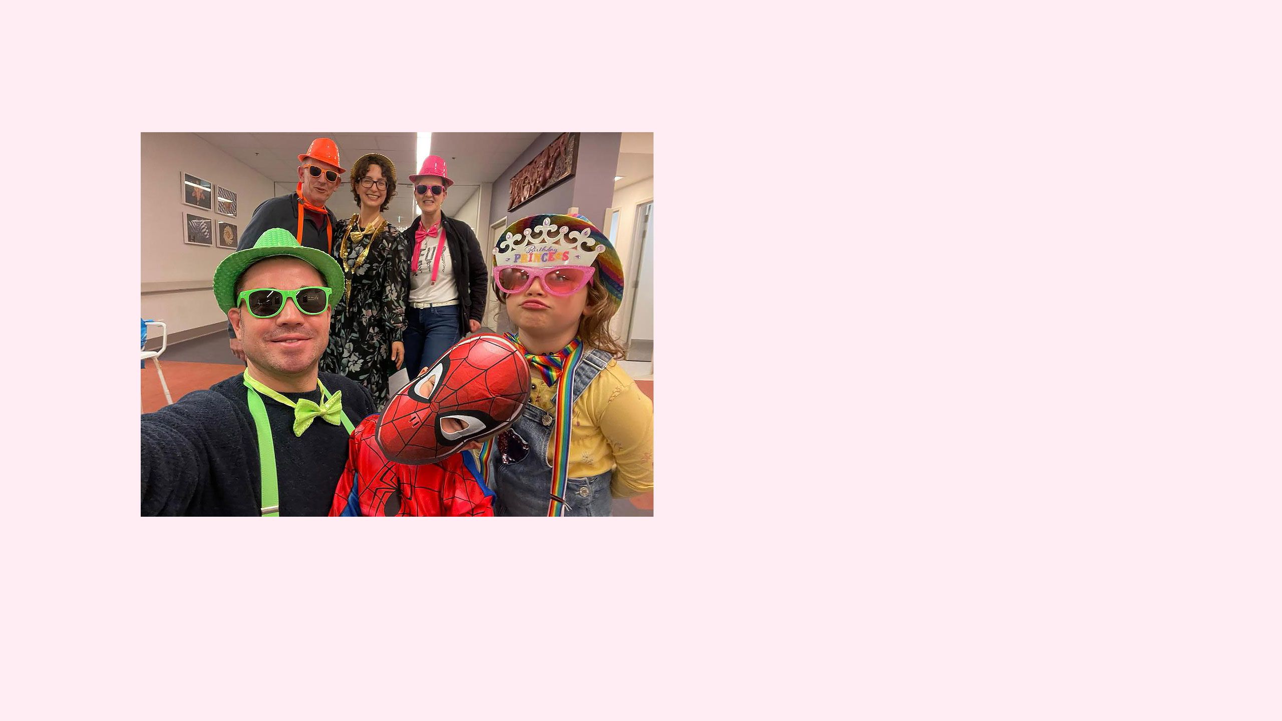 Lauren’s husband Antony is in the front-left corner, wearing a green hat, green bowtie and green glasses. He is standing next to Alfie, dressed as Spiderman, who is next to Evangeline, wearing a rainbow hat and suspenders, pink glasses and a ‘Birthday Princess’ tiara. In the background, Lauren is in the middle of two friends, wearing a gold hat, bowtie and necklace.