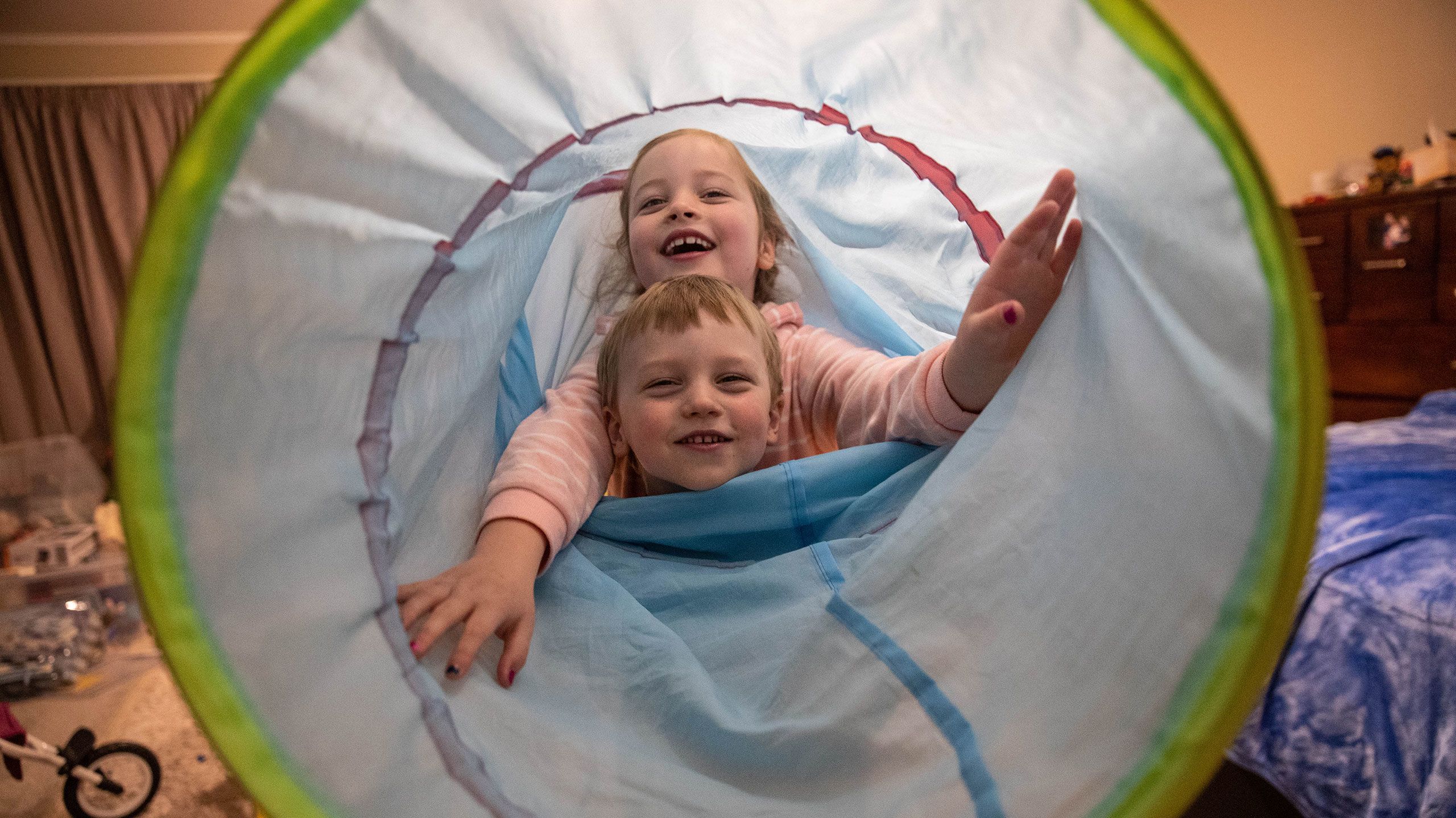 Alfie and Evangeline are smiling from inside a fabric play tunnel.